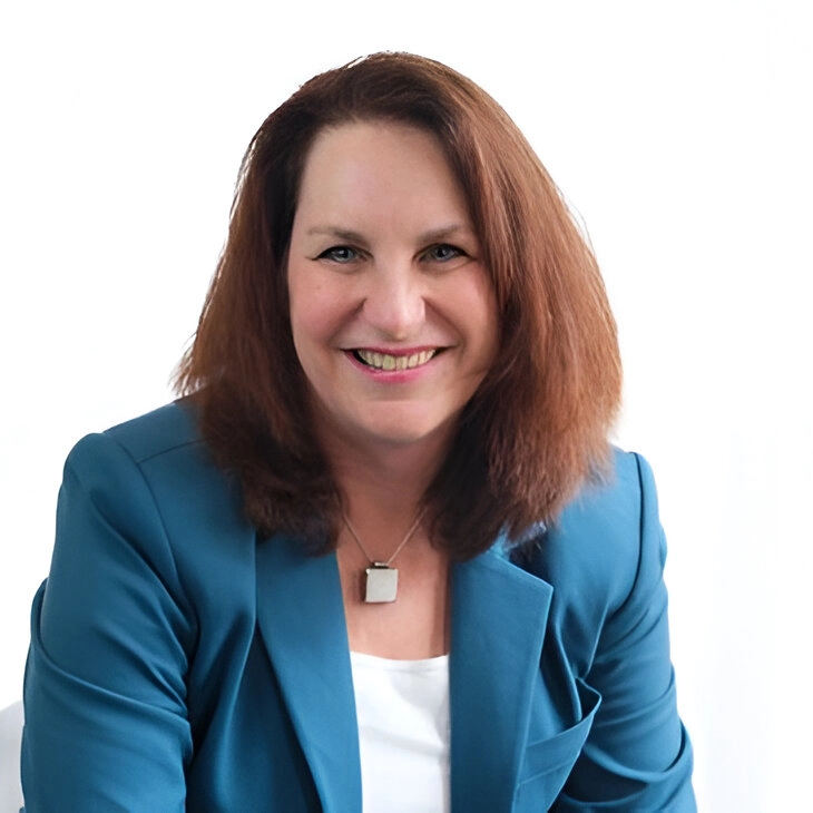 A person with shoulder-length brown hair wearing a teal blazer and white shirt smiles against a plain white background.