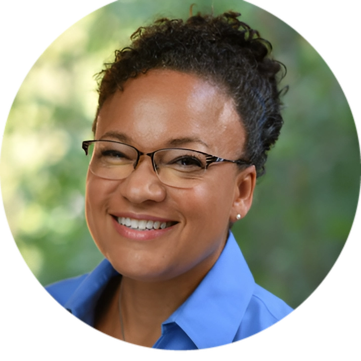 Person with curly hair and glasses smiling, wearing a blue shirt against a blurred green background.